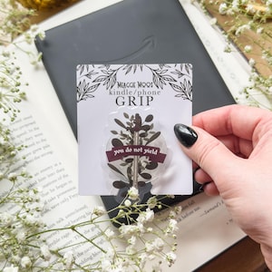 May include: A clear phone grip with a botanical design and the text "you do not yield" is held over a Kindle. The grip is on a card that says "MEAGGIE MOOS kindle/phone GRIP". The background includes a book and white flowers.