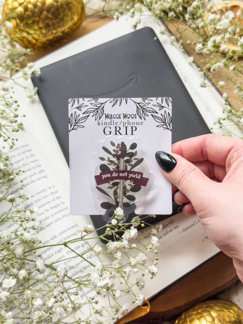 May include: A clear phone grip with a botanical design and the text "you do not yield" is held over a Kindle. The grip is on a card that says "MEAGGIE MOOS kindle/phone GRIP". The background includes a book and white flowers.