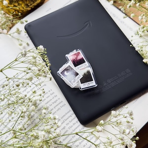 May include: A black Kindle e-reader with a clear sticker featuring three small landscape images and text. The e-reader rests on an open book with white flowers and a gold decorative sphere. The sticker images include text that reads "Crescent City", "Twilight", and "Forest".