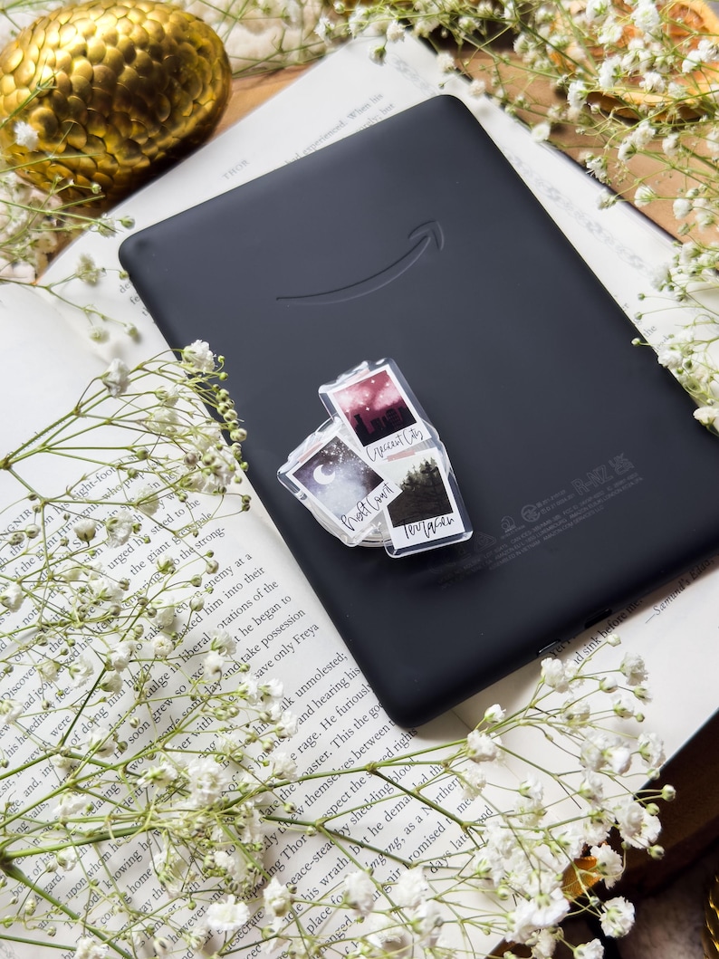 May include: A black Kindle e-reader with a clear sticker featuring three small landscape images and text. The e-reader rests on an open book with white flowers and a gold decorative sphere. The sticker images include text that reads "Crescent City", "Twilight", and "Forest".