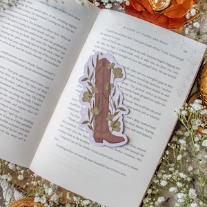 Puede incluir: Un marcapáginas con un diseño de bota de vaquero marrón con hojas y tallos verdes, sobre un fondo lila claro. El marcapáginas está colocado en un libro abierto.