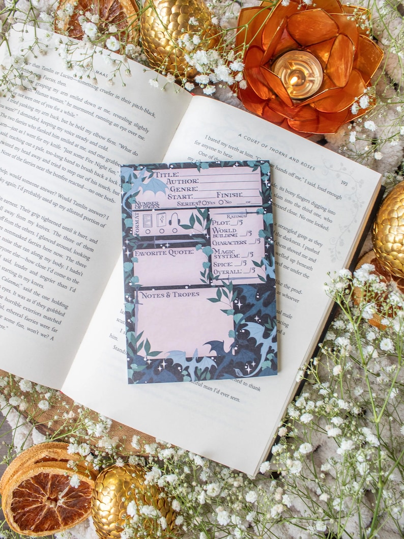 May include: A book with a bookmark featuring a floral and dragon design, with text fields for notes. The book is open to a page with text, surrounded by flowers and decorative elements.