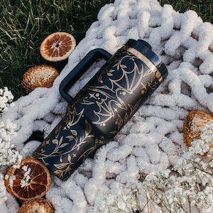 May include: A black travel mug with a gold design of bats and vines. The mug has a black handle and a gold lid. The mug is sitting on a white fluffy blanket.