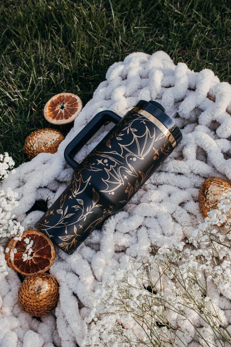 May include: A black travel mug with a gold design of bats and vines. The mug has a black handle and a gold lid. The mug is sitting on a white fluffy blanket.
