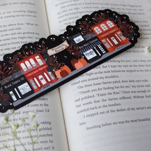 May include: A black and red illustrated bookmark with a Halloween theme. The bookmark features a row of shops with names like "Witches Brew", "Boo Sheet Company", and "Haunted Library". The shops are decorated with pumpkins, bats, and stars.