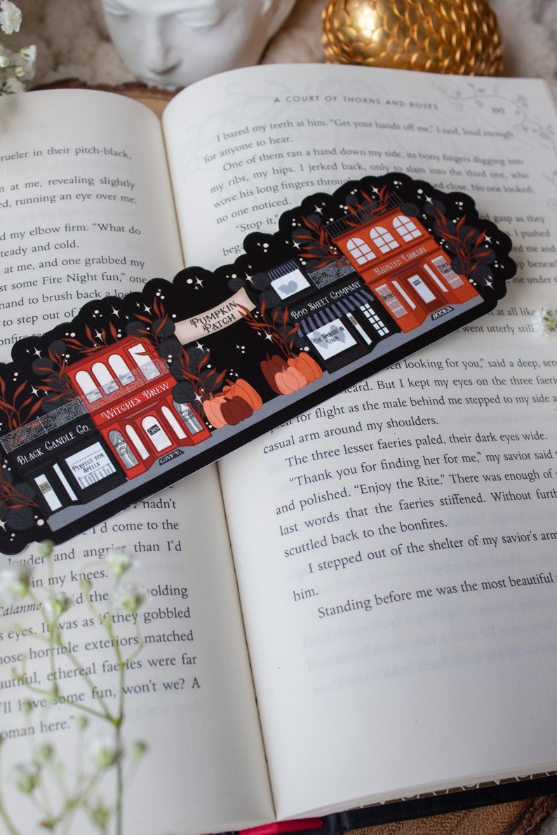 May include: A black and red illustrated bookmark with a Halloween theme. The bookmark features a row of shops with names like "Witches Brew", "Boo Sheet Company", and "Haunted Library". The shops are decorated with pumpkins, bats, and stars.