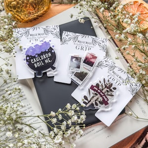 May include: A collection of phone grips with various designs, including a cauldron with the text "CAULDRON BOIL ME," a cityscape, and botanical elements. The grips are displayed on a book with white flowers and a golden sphere.