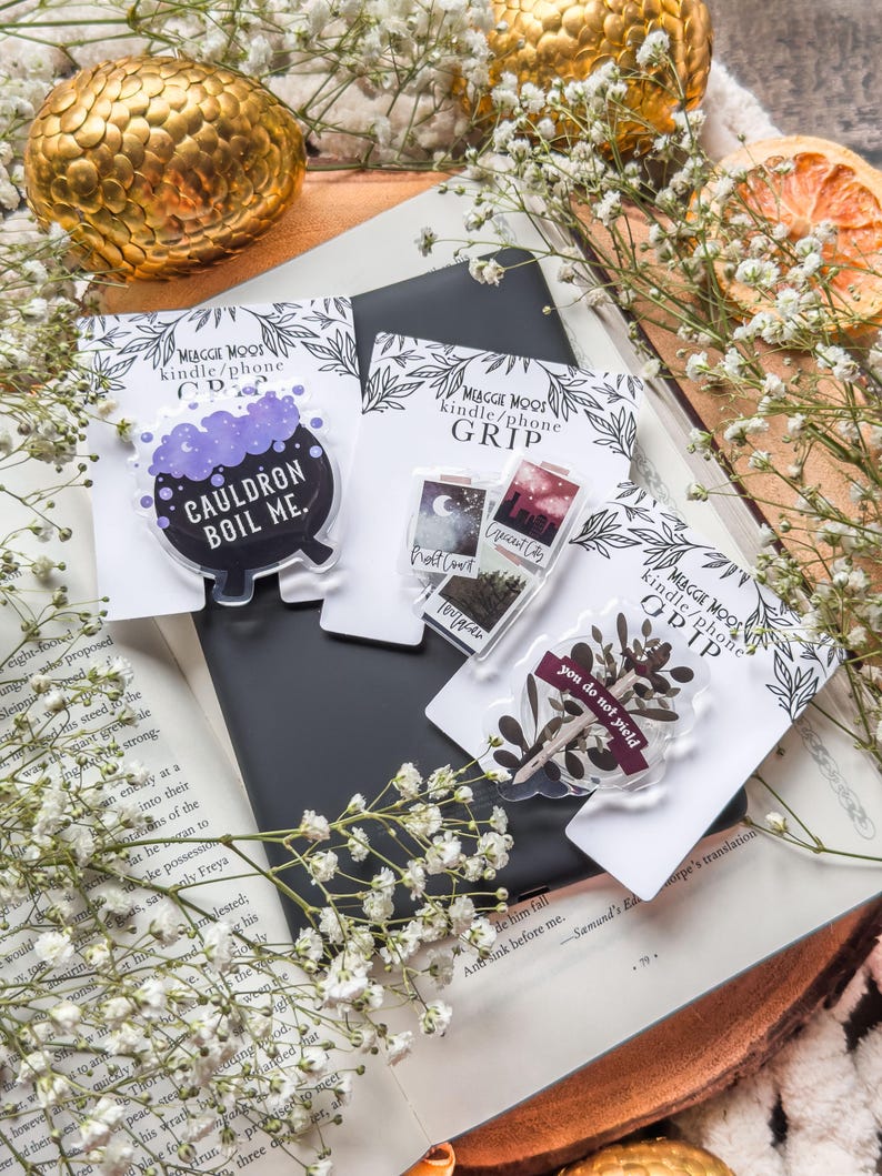 May include: A collection of phone grips with various designs, including a cauldron with the text "CAULDRON BOIL ME," a cityscape, and botanical elements. The grips are displayed on a book with white flowers and a golden sphere.