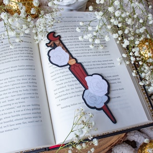 May include: A decorative bookmark featuring a red and black design with white rose accents. The bookmark is placed on an open book with white flowers and a candle in the background, creating a romantic aesthetic.