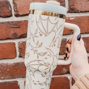 May include: White stainless steel travel mug with a gold rim and a handle. The mug has a floral and bat design in gold.