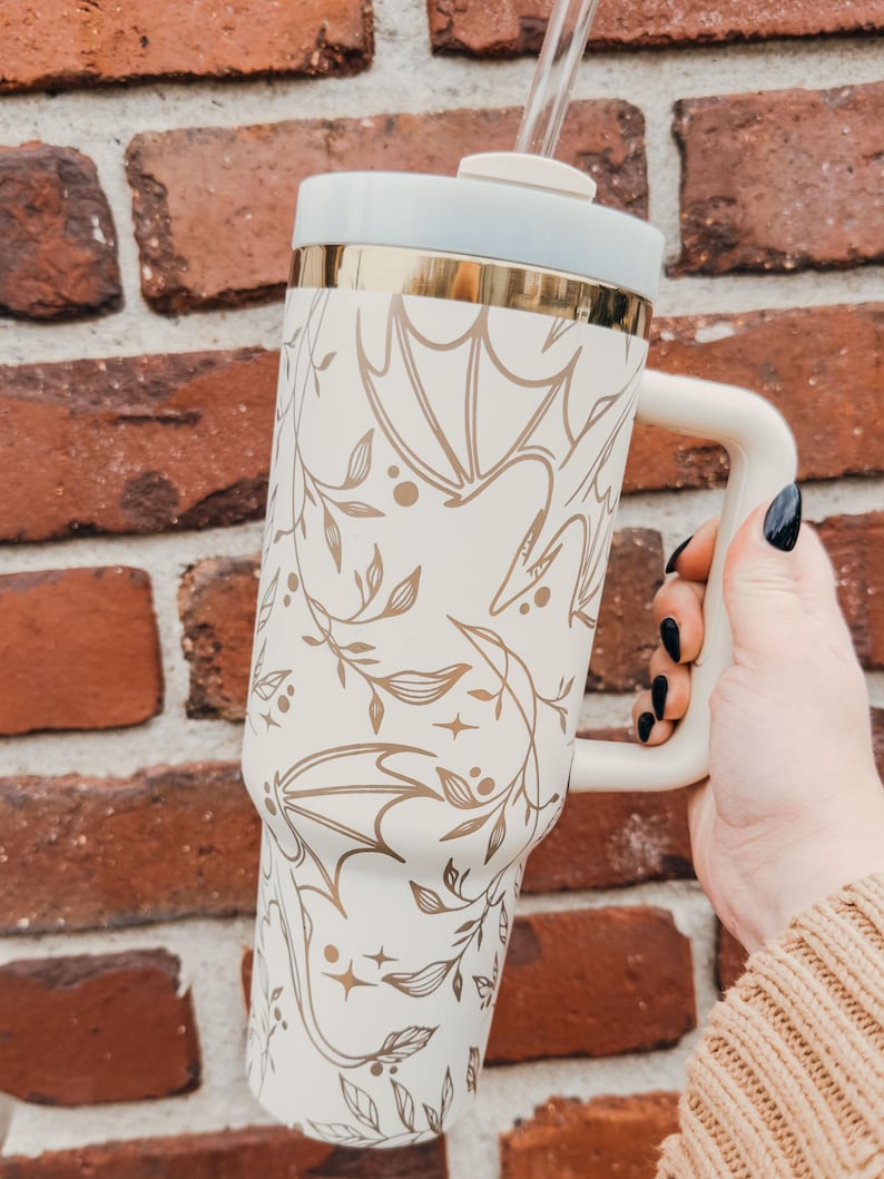 May include: White stainless steel travel mug with a gold rim and a handle. The mug has a floral and bat design in gold.