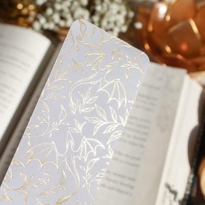 May include: A white bookmark with a gold dragon and leaf design. The bookmark is rectangular and features a repeating pattern of dragons and leaves. The design is in gold foil, creating a shimmering effect. The bookmark is placed in an open book.