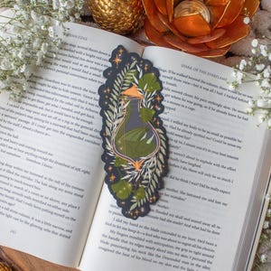 Puede incluir: Marcapáginas decorativo negro con una ilustración de una botella de poción con líquido verde y elementos botánicos. El marcapáginas tiene una forma única y está sobre un libro abierto con texto.