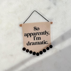 May include: A small banner with the text "So apparently, I'm dramatic." The banner is beige with black text and black pom-poms along the bottom. It has a wooden dowel and a black string for hanging.