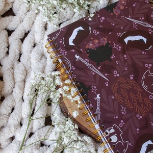Puede incluir: Una libreta de espiral color burdeos con un dise&ntilde;o de Halloween espeluznante que presenta murci&eacute;lagos, calderos y ata&uacute;des. La libreta est&aacute; sobre una superficie blanca y peluda.