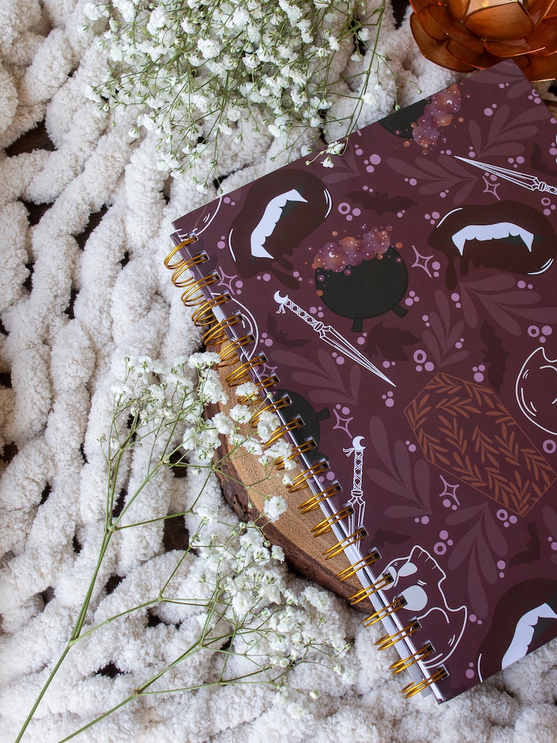 Puede incluir: Una libreta de espiral color burdeos con un dise&ntilde;o de Halloween espeluznante que presenta murci&eacute;lagos, calderos y ata&uacute;des. La libreta est&aacute; sobre una superficie blanca y peluda.
