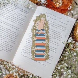 May include: A decorative bookmark featuring a stylized illustration of a figure atop a stack of books, framed by a leafy border. The bookmark has orange, blue, and white accents. The bookmark is placed on the open pages of a book.