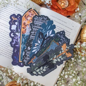 Pu&ograve; includere: Tre segnalibri con uno schema di colori blu scuro e verde che rappresentano un drago nero, una pila di libri e un drago con le ali spiegate. I segnalibri sono su un libro aperto con il testo "A Court of Thorns and Roses" visibile.