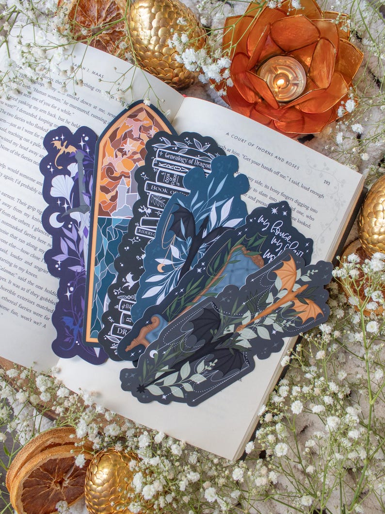 Pu&ograve; includere: Tre segnalibri con uno schema di colori blu scuro e verde che rappresentano un drago nero, una pila di libri e un drago con le ali spiegate. I segnalibri sono su un libro aperto con il testo "A Court of Thorns and Roses" visibile.