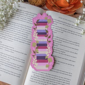 Peut inclure: Marque-page rose avec une pile de livres violets, roses et blancs, orné de feuilles vertes. Le marque-page est posé sur un livre ouvert avec du texte.
