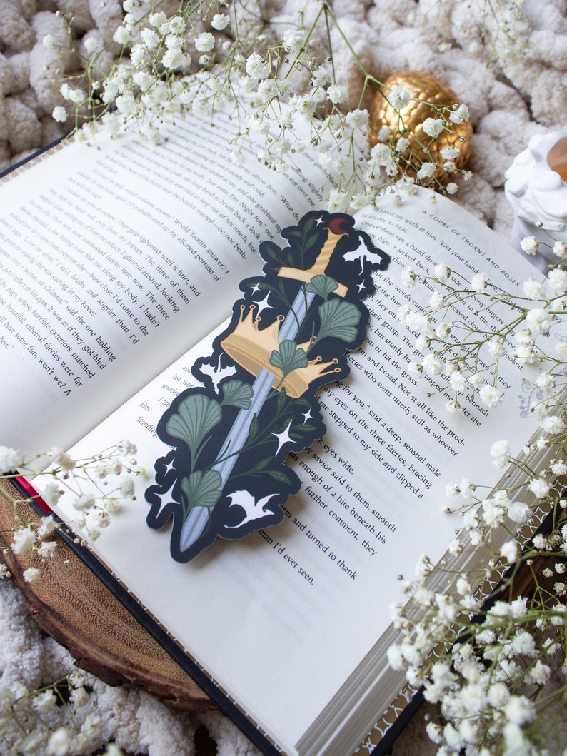 May include: A bookmark featuring a silver sword with a gold crown and green leaves on a black background. The bookmark is a decorative item for book lovers.