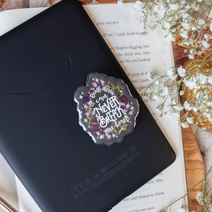 May include: A black e-reader with a clear sticker that reads "Dead lovers can never break your heart" in white script, surrounded by a floral design. The e-reader rests on an open book with white flowers.