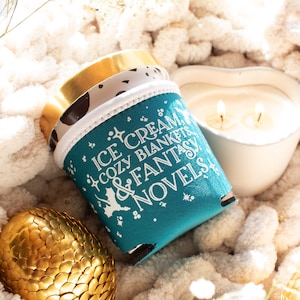 May include: A teal drink sleeve with the text "Ice Cream, Cozy Blankets & Fantasy Novels" sits next to a heart-shaped candle and a golden decorative egg. The sleeve has a white trim and a gold rim.