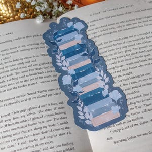 May include: A blue and white bookmark with a stack of books design. The bookmark features a floral and leaf pattern, with a light blue background and a stack of books in shades of blue and pink. The bookmark is on top of an open book.