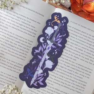 May include: A purple and white bookmark featuring a sword design with floral and celestial accents. The bookmark has a dragon silhouette and a crescent moon, perfect for fantasy book lovers.