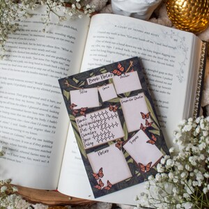 K&ouml;nnte beinhalten: Ein offenes Buch, eine dekorative Kerze und eine goldene Kugel sind mit einer Notizkarte f&uuml;r B&uuml;cher angeordnet. Die Karte enth&auml;lt Felder f&uuml;r Titel, Genre, Autor und Lieblingszitat mit Schmetterlingsillustrationen.