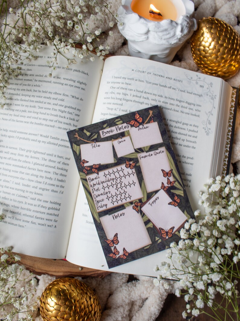 K&ouml;nnte beinhalten: Ein offenes Buch, eine dekorative Kerze und eine goldene Kugel sind mit einer Notizkarte f&uuml;r B&uuml;cher angeordnet. Die Karte enth&auml;lt Felder f&uuml;r Titel, Genre, Autor und Lieblingszitat mit Schmetterlingsillustrationen.