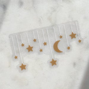 May include: A clear sticker featuring a gold crescent moon and stars hanging from white lines. The stars vary in size and are evenly spaced. The sticker is on a white and gray marble surface.