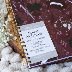 Puede incluir: Un cuaderno espiral morado con un dise&ntilde;o espeluznante que presenta calderos, murci&eacute;lagos y colmillos. El cuaderno mide 21,6 cm por 27,9 cm y tiene 100 hojas de papel de doble cara con l&iacute;neas de universidad. El texto en la portada dice "Spiral Notebook" y "meaggiemoos.com".
