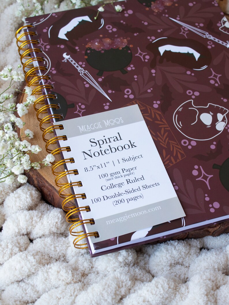 Puede incluir: Un cuaderno espiral morado con un dise&ntilde;o espeluznante que presenta calderos, murci&eacute;lagos y colmillos. El cuaderno mide 21,6 cm por 27,9 cm y tiene 100 hojas de papel de doble cara con l&iacute;neas de universidad. El texto en la portada dice "Spiral Notebook" y "meaggiemoos.com".