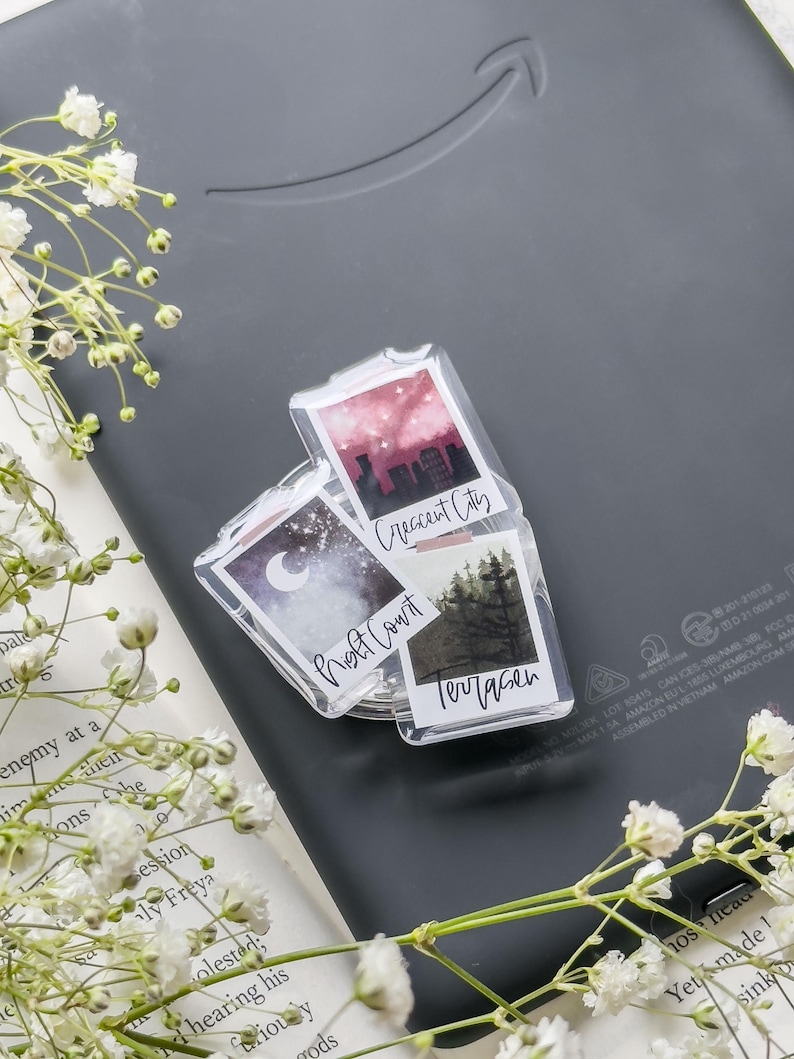 May include: Clear acrylic Polaroid-style magnets with landscape art and text. The magnets feature images of a night sky, a cityscape, and a forest. The text includes "Night Court", "Crescent City", and "Terrasen".