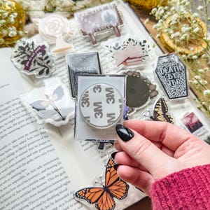 May include: A collection of clear acrylic stickers with various designs, including a butterfly, a sign that says "The City of Forms Welcomes You", and the text "Death by TBR". The stickers are displayed on an open book.