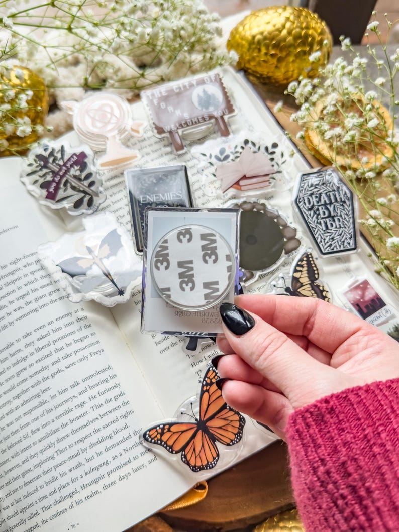 May include: A collection of clear acrylic stickers with various designs, including a butterfly, a sign that says "The City of Forms Welcomes You", and the text "Death by TBR". The stickers are displayed on an open book.