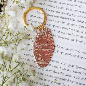 Puede incluir: Un llavero dorado con un colgante ovalado de color marr&oacute;n rojizo. El colgante presenta el texto "Keys to my Haunted Library, Readers Only" con ilustraciones bot&aacute;nicas y de libros. Flores blancas rodean el llavero.