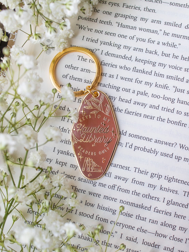 Puede incluir: Un llavero dorado con un colgante ovalado de color marr&oacute;n rojizo. El colgante presenta el texto "Keys to my Haunted Library, Readers Only" con ilustraciones bot&aacute;nicas y de libros. Flores blancas rodean el llavero.