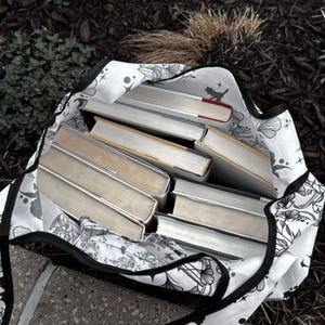 Peut inclure: Un sac fourre-tout blanc avec un motif floral noir et une fermeture &eacute;clair noire. Le sac est rempli de plusieurs livres reli&eacute;s.