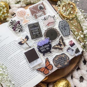 May include: A collection of clear acrylic charms and stickers with various designs, including butterflies, a cauldron, and text that reads "Enemies to Lovers" and "The City of Forks Welcomes You." The items are arranged on an open book.