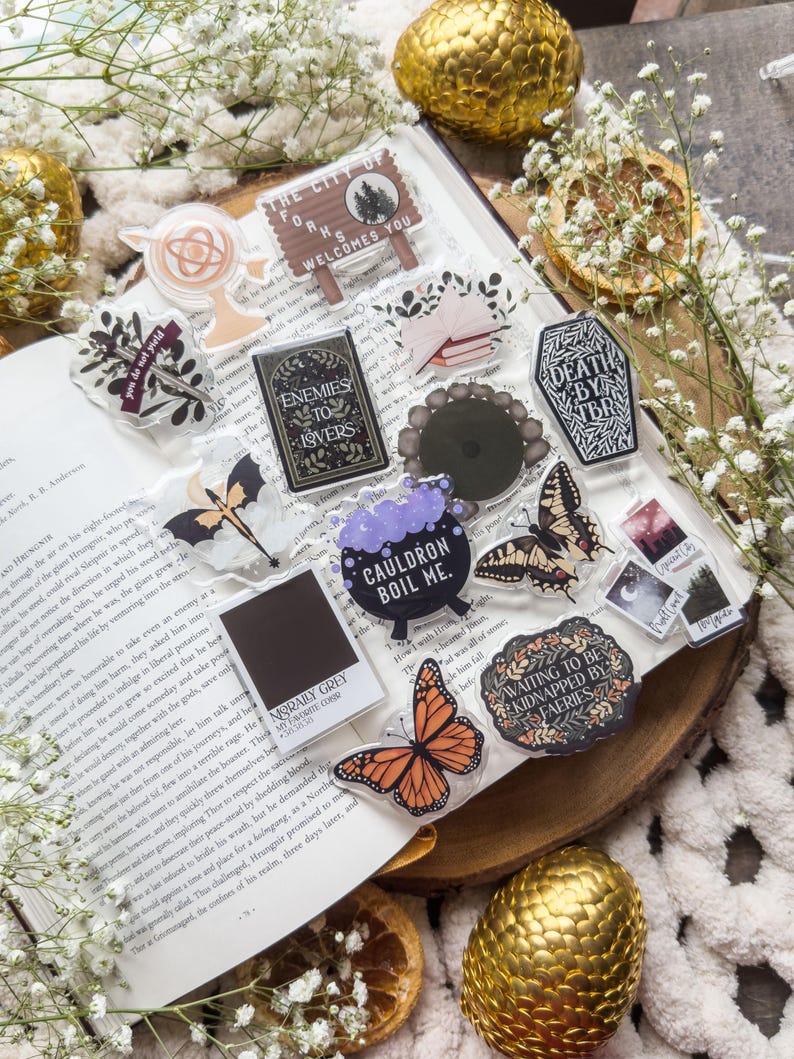 May include: A collection of clear acrylic charms and stickers with various designs, including butterflies, a cauldron, and text that reads "Enemies to Lovers" and "The City of Forks Welcomes You." The items are arranged on an open book.