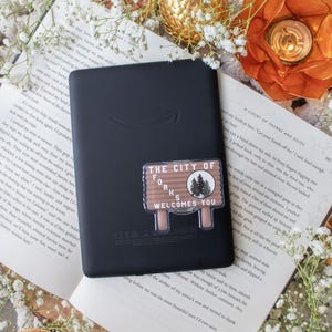 May include: Black e-reader with a sticker that reads "THE CITY OF FORKS WELCOMES YOU." The sticker is brown and white with a forest graphic. The e-reader is on top of an open book with white flowers and orange slices.
