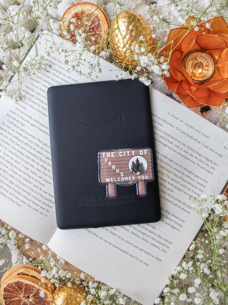 May include: Black e-reader with a sticker that reads "THE CITY OF FORKS WELCOMES YOU." The sticker is brown and white with a forest graphic. The e-reader is on top of an open book with white flowers and orange slices.