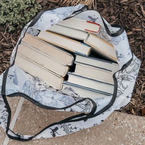 Peut inclure: Un sac fourre-tout blanc avec un motif floral noir et une bordure noire. Le sac est rempli d'une pile de livres broch&eacute;s.
