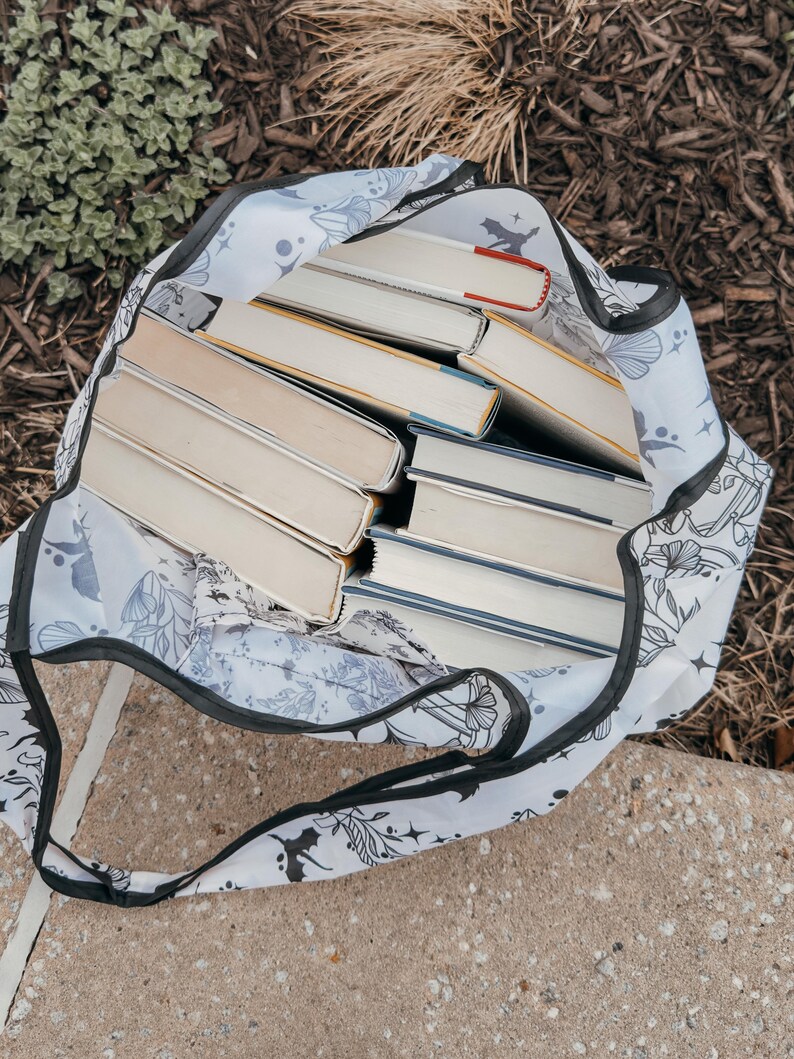 Peut inclure: Un sac fourre-tout blanc avec un motif floral noir et une bordure noire. Le sac est rempli d'une pile de livres broch&eacute;s.