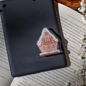 May include: A black e-reader with a gingerbread house sticker. The sticker is white with orange accents and the word "LIBRARY" on it. The e-reader is resting on an open book with white flowers and a candle in the background.