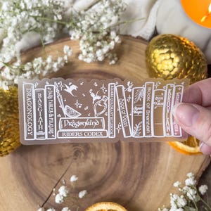 May include: A clear sticker featuring a bookshelf design with titles like "Dragonology" and "Riders Codex." The sticker includes illustrations of a sword, stars, and a dragon. The sticker is held against a wooden surface with dried orange slices and white flowers.
