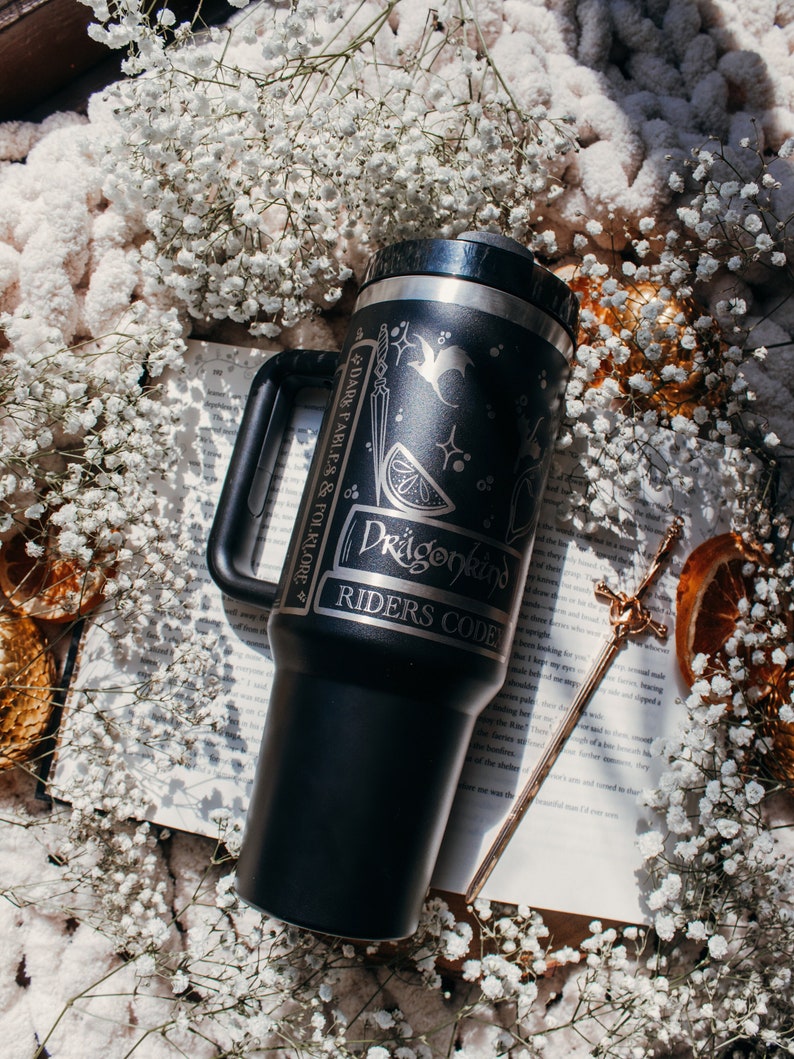 May include: Black stainless steel travel mug with a silver handle. The mug has a black and white design with a dragon, a sword, and a lemon. The text "Dragonkind Riders Code" is engraved on the mug.