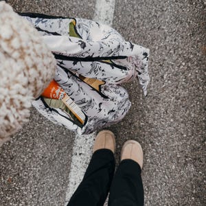 Peut inclure: Une personne portant un pantalon noir et un pull blanc duveteux tient un sac de courses r&eacute;utilisable blanc avec un motif floral noir et blanc. Le sac est rempli d'articles, dont une bo&icirc;te de craquelins. La personne se tient sur une surface d'asphalte grise.